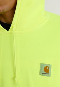 Bright yellow hoodie with a soft texture, featuring a small square logo patch in green and orange on the front. Drawstring hood details visible.