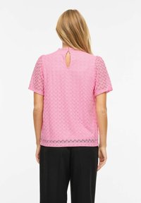 Haut rose à manches courtes texturé avec un détail dos en forme de goutte, terminé par une bordure en dentelle noire à l'ourlet, associé à un pantalon noir.