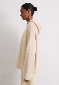 Sweat à capuche en tricot beige, oversized, avec un design texturé, présentant une poche avant et des poignets côtelés. Le tissu semble doux et chaud.
