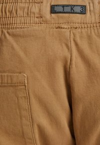 Khaki bavlněné kalhoty s nařaseným pasem, boční kapsou a černou značkovou nášivkou LTK3 blízko horní části zad.