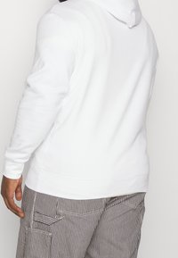 Sudadera blanca hecha de tela suave, con mangas largas y capucha con cordón, combinada con pantalones cargo a rayas.