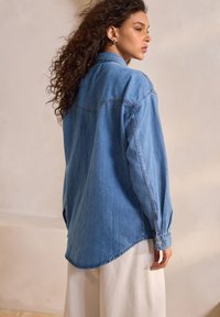 Camicia di jeans azzurro chiaro con maniche lunghe, vestibilità ampia, orlo curvo, polsini con bottoni e dettagli di cucitura minima sulla schiena.