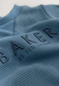 Modrá textilná tkanina s vaflovým vzorom, s vyšitým logom "BAKER" v tmavšej modrej, umiestneným vpredu.