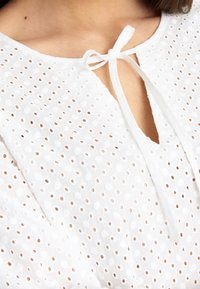 Top blanco con un patrón de ojales texturizado, escote en forma de llave con detalle de lazada y mangas cortas. Material suave y ligero.