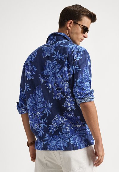 Άντρας με μακρυμάνικο πουκάμισο σκούρου μπλε με λουλούδια και μπεζ παντελόνι, γυρισμένος πλάγια, φοράει γυαλιά ηλίου και έχει τα μανίκια σηκωμένα.