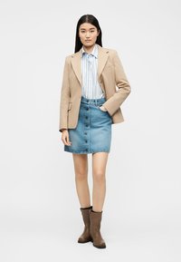 Γυναίκα που φοράει μπεζ σακάκι, μπλε ριγέ πουκάμισο, φούστα από τζιν με κουμπιά και καφέ μποτάκια μέχρι τον αστράγαλο, στέκεται με το ένα χέρι στην τσέπη.