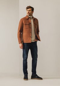 Bruine wollen jas met twee borstzakken, knoopsluiting en een kraagontwerp, gedragen over een lichtgekleurde coltrui en donkere jeans.