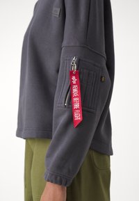 Šedá mikina s volným střihem, s kapsou na zip na rukávu a červenou visačkou s nápisem „REMOVE BEFORE FLIGHT“. Texturovaná látka.