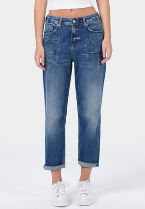 Frau trägt mittelhohe, locker sitzende blaue Jeans mit aufgekrempelten Säumen, aufgesetzten Taschen und weißen Sneakers, dargestellt von der Taille bis zu den Füßen vor weißem Hintergrund.