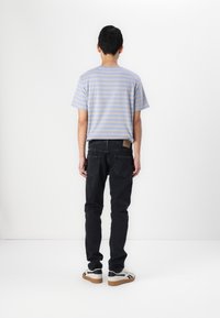 T-shirt rayé bleu et beige avec une coupe décontractée, associé à un jean noir et des baskets blanches avec des accents noirs et marron, vu de dos.