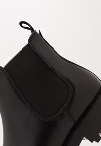 Botín Chelsea de cuero negro con paneles laterales elásticos, suela de goma resistente y tirador para facilitar el uso. Textura suave con acentos de costura.