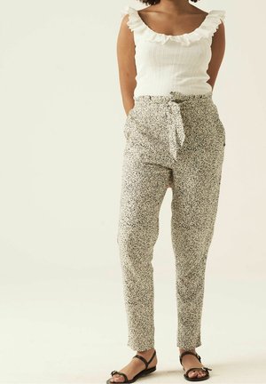 Vrouw draagt een witte mouwloze top met ruches, hooggesneden gebloemde broek met ceintuur, en zwarte platte sandalen, staand met haar handen achter haar rug.