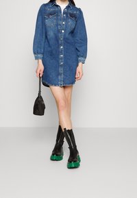 Femme portant une robe en jean bleue boutonnée, tenant un petit sac à main noir et des bottes noires avec des semelles épaisses vertes.