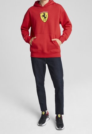Persoon die een rode hoodie draagt met een geel Ferrari-logo, donkere spijkerbroek en zwarte sneakers met bijpassende gele Ferrari-logo’s.