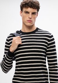Suéter de rayas en blanco y negro con cuello redondo acanalado y mangas largas. El modelo sostiene una correa negra, mostrando un diseño ajustado.