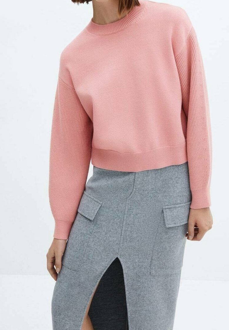 Femme portant un pull rose à manches longues et une jupe midi grise avec une fente avant et des poches à rabat, sur un fond uni.