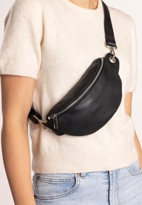 Black leather fanny pack with a curved design, silver zipper, and adjustable strap, worn across the body over a light sweater.