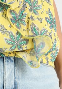 Gul blus med blommönster och volanger; har mönster av gröna och grå blad; tillverkad av lätt, genomskinlig tyg. Kombinerad med denimshorts.