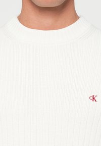 Pulover alb din tricot cu dungi, cu guler rotund și un mic logo roșu "CK" pe pieptul stâng. Țesătura texturată oferă un aspect moale.