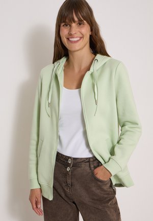 Femme souriante portant un sweat à capuche zippé vert clair, un débardeur blanc et un pantalon marron, la main dans la poche, sur fond uni.