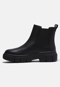 Botte Chelsea en cuir noir avec semelle en caoutchouc épaisse, panneaux latéraux élastiques et languette à l’arrière, montrée de profil sur fond clair.