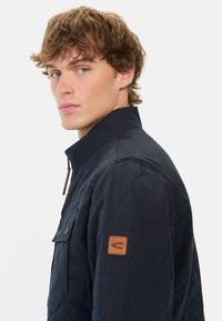 Granatowa pikowana kurtka z wysokim kołnierzem, wyposażona w kieszeń na piersi, zatrzaski oraz skórzany akcent na rękawie.