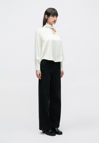 Biała bluzka z długim rękawem, wysokim kołnierzem i ozdobnym wycięciem na przodzie, zestawiona z czarnymi, szerokimi spodniami i czarnymi butami na platformie.