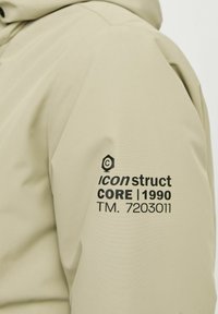 Beige vandtæt jakke med hætte, glat tekstur og sort logo-detalje på venstre ærme med tekst elementer.