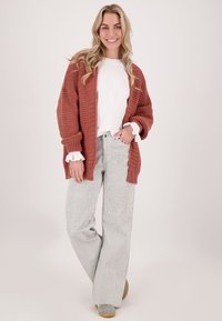 Roestkleurige gebreide cardigan over een witte langeketelshirt, gecombineerd met grijs gestreepte wijde broek en grijze instapschoenen.