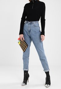 Pull à col zippé noir, jean bleu clair taille haute et bottines à talons noires avec bouts argent és. Tenant une pochette rayée.