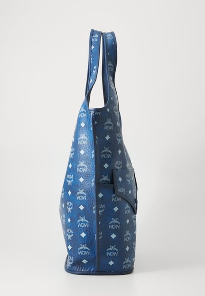 Μπλε δερμάτινη τσάντα tote με επαναλαμβανόμενα λευκά λογότυπα MCM, διαμαντένια μοτίβα και μπροστινή τσέπη, εμφανίζεται από το πλάι σε λευκό φόντο.