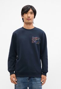 Sweatshirt bleu marine en coton doux, avec un col rond et un motif graphique multicolore brodé sur le côté gauche de la poitrine. Coupe décontractée.