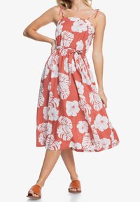 Mujer vistiendo un vestido sin mangas de color coral con estampado de flores y hojas tropicales en blanco, atado a la cintura, combinado con sandalias deslizadas marrones.