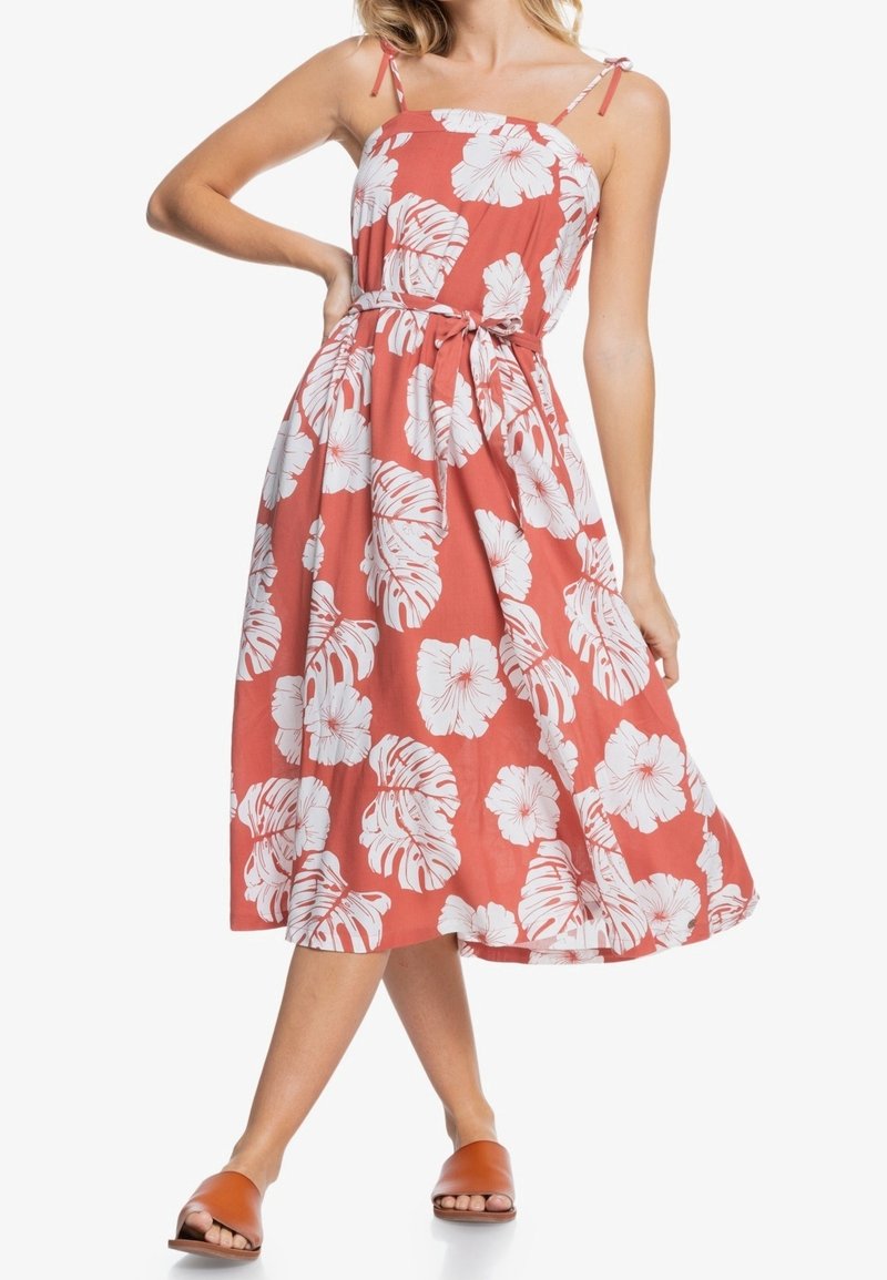 Mujer vistiendo un vestido sin mangas de color coral con estampado de flores y hojas tropicales en blanco, atado a la cintura, combinado con sandalias deslizadas marrones.