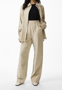 Woman wearing beige wide-leg pants with elastic waist, matching beige open button-up shirt, black top, and beige square-toe shoes.