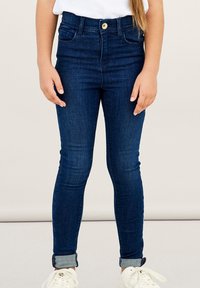 Vaqueros ajustados de denim azul oscuro, con cierre de botón, dos bolsillos frontales y puños enrollados en los tobillos.