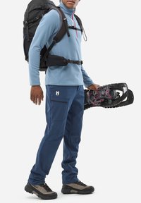 Pull en polaire bleue avec zip, pantalon de randonnée bleu marine avec poches, sac à dos noir et raquettes à neige. Chaussures de randonnée grises et noires avec lacets.