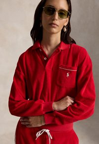 Femme aux cheveux foncés portant des lunettes de soleil à verres verts, un polo rouge à manches longues et un pantalon rouge assorti avec un cordon blanc.