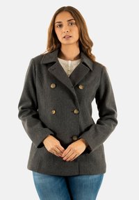 Cappotto grigio in lana doppiopetto con colletto a punte, caratterizzato da due file di bottoni circolari e tasche frontali. Indossato sopra un top leggero.