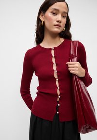 Burgundi spalvos susiglaudęs kardiganas su perlų mygtukais ir unikaliu išpjoviniu priekyje. Suderintas su blizgiu burgundiškos spalvos rankine.