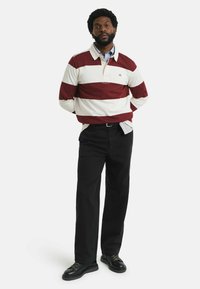 Rot-weiß gestreiftes Rugby-Shirt mit Kragen, über einem hellblauen Hemd mit Knöpfen. Schwarze Hosen und Schnürschuhe.