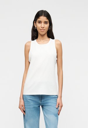 Fiatal nő, hosszú sötét hajjal, ujjatlan fehér felsőt és világoskék farmert visel, egyszínű világos háttér előtt áll.