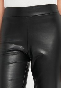 Pantalons en simili cuir noir avec une texture lisse et une taille haute, présentant des coutures décoratives et une silhouette ajustée.