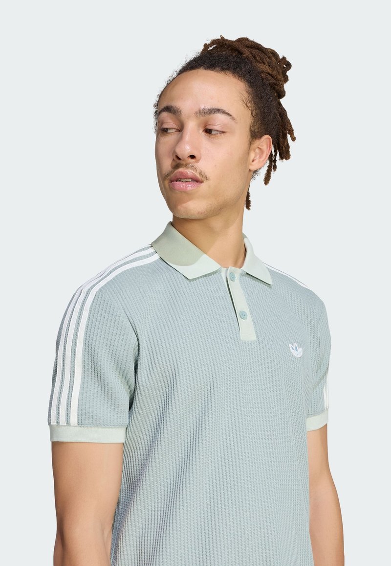 Mann mit Dreadlocks, der ein hellgrünes, strukturiertes Poloshirt mit weißen Streifen an den Ärmeln trägt und nach links blickt, vor einem einfarbigen Hintergrund.