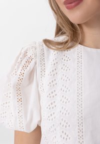 Biała bluzka z bufkami, złożonymi detalami w formie wycięć oraz haftów z przodu. Gładka tekstura i luźny krój.