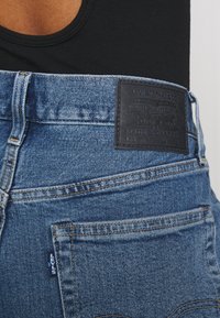 Jeans de mezclilla azul con un parche de cuero en la cintura que presenta el logo "Levi Strauss & Co.", costuras naranjas y una pequeña etiqueta de marca en el bolsillo trasero.