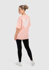 Camiseta oversized rosa claro con gráfico de mariposa blanca en la parte posterior, combinada con leggings negros y zapatillas blancas. Textura de tela suave.