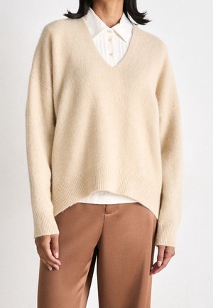Maglione beige a scollo a V realizzato in materiale morbido, con orlo e polsini a coste, indossato sopra una camicia bianca con colletto e abbinato a pantaloni marroni.