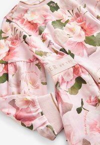 Tissu rose doux avec de grandes roses style aquarelle et des feuilles vertes, poignet froncé et une étiquette de marque cousue.