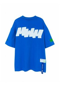 Camiseta de algodón azul con logo gráfico blanco oversize, mangas cortas, escote redondo y una etiqueta de acento verde en la manga izquierda.